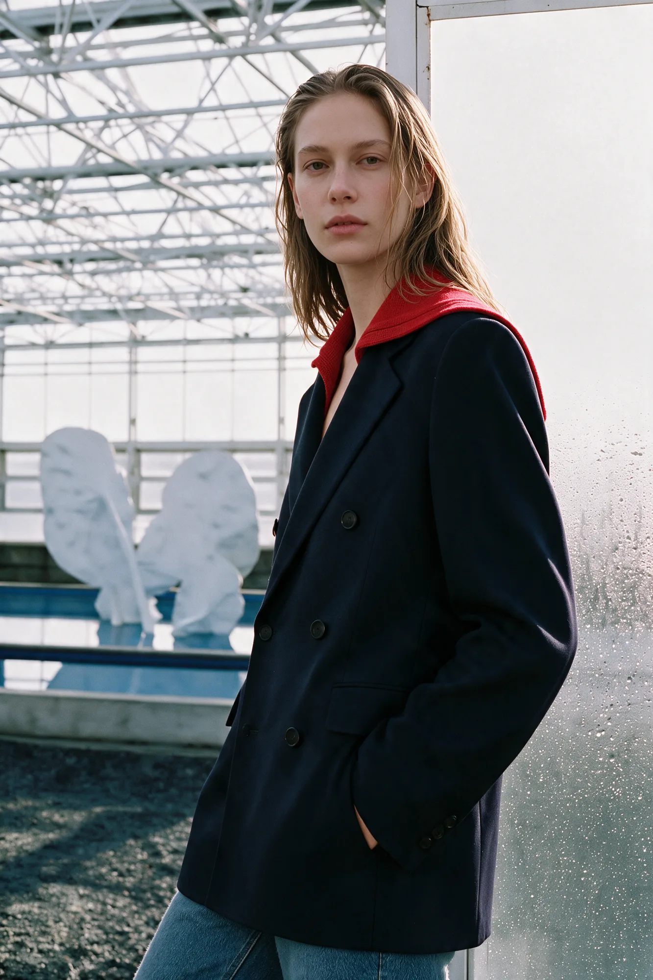 Model in a dark tailored jacket standing inside a sunlit greenhouse